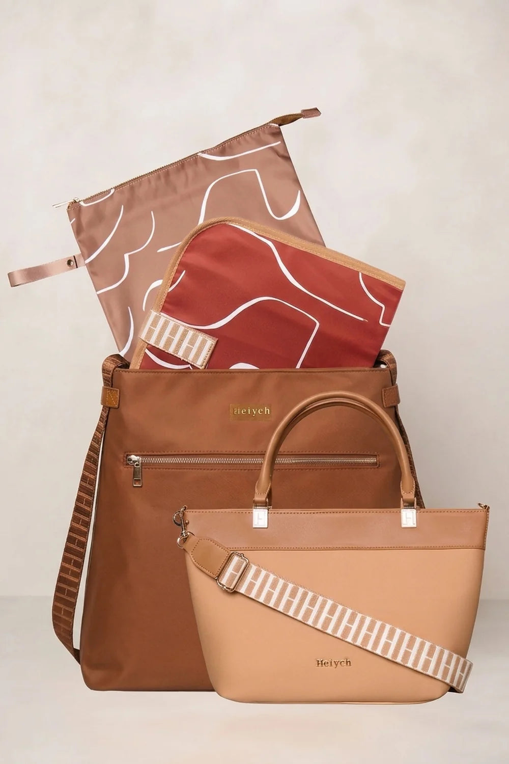 A coordinated travel bag set from Heyych, featuring a cognac tan tote bag, a rust-red zippered pouch with white abstract lines, and a dusty-rose reusable bag with a white line pattern, all arranged on a light beige background.