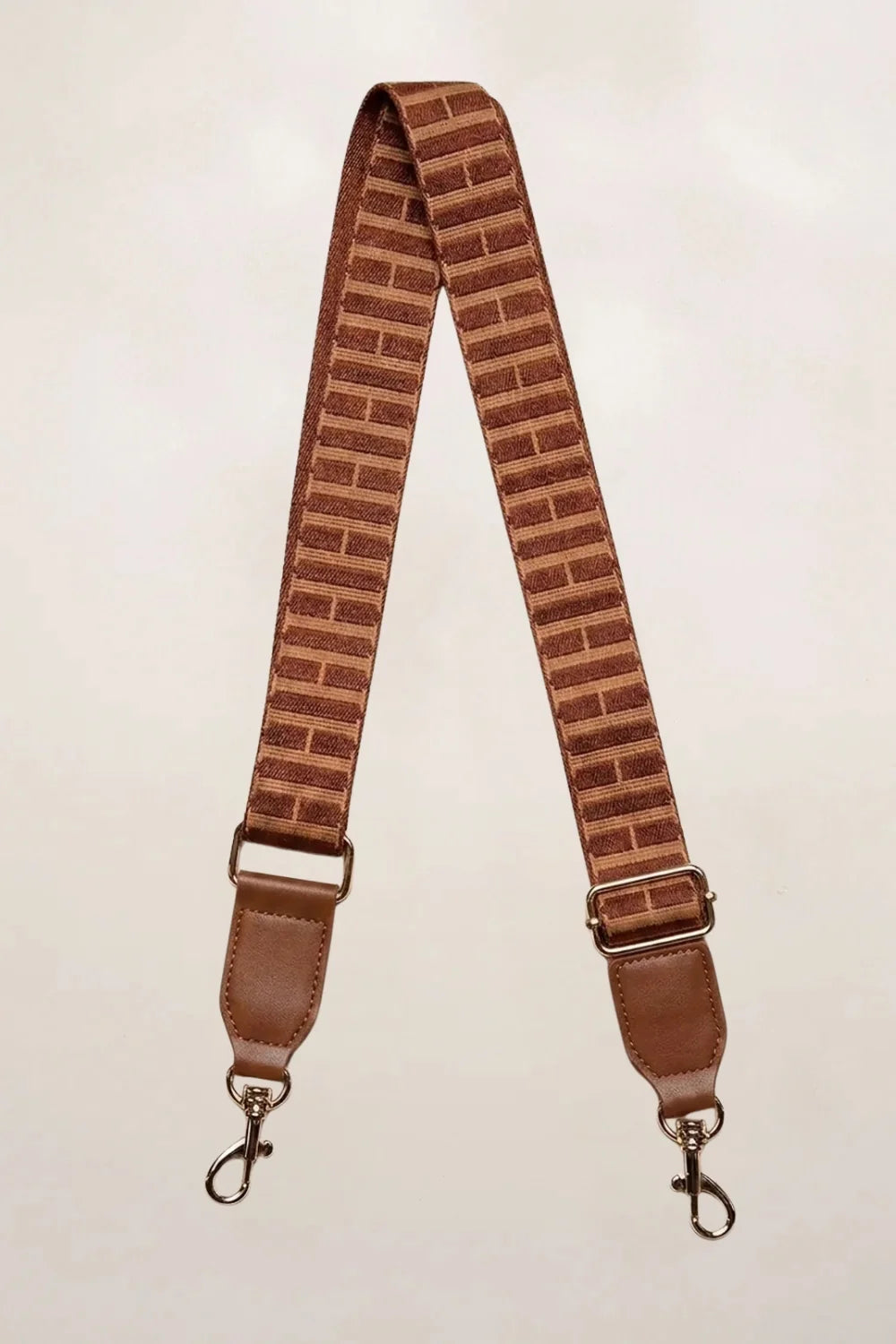 A cognac fabric shoulder strap with a brick-patterned design and brown leather-like ends, featuring metal swivel clips on a light beige background.