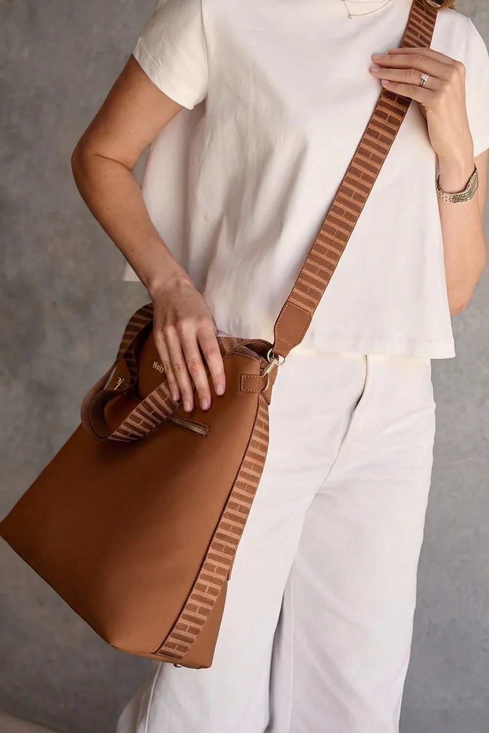 A woman wearing a white short-sleeve top and white pants carries a cognac tan crossbody bag with a wide brown-and-white striped strap against a gray wall.